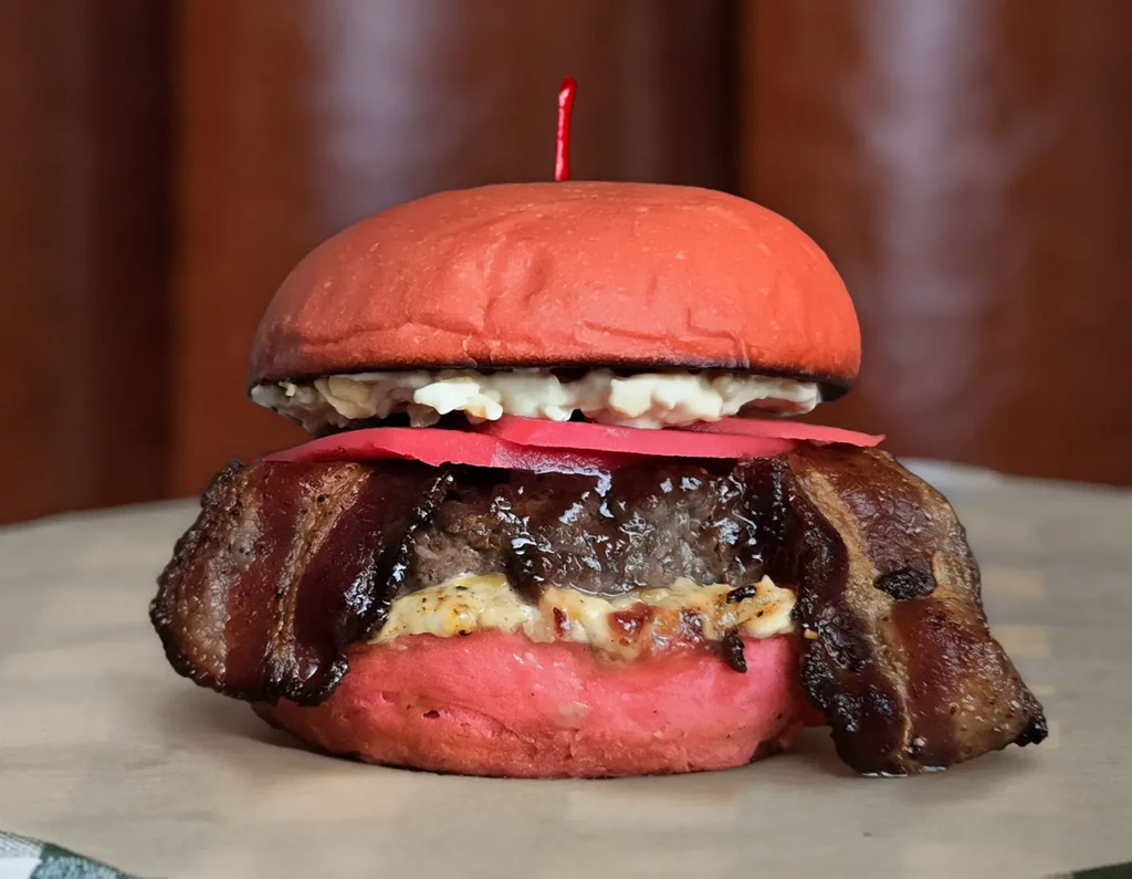 Close-up of a burger with a red bun, bacon, and coleslaw.