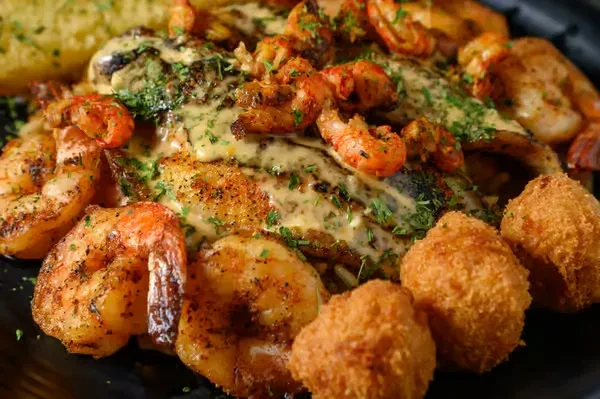 Grilled fish fillets, shrimp, and hushpuppies on a dark plate.