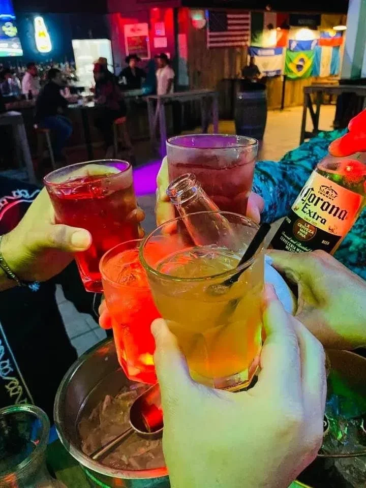 Hands toast drinks in a lively, dimly lit bar.