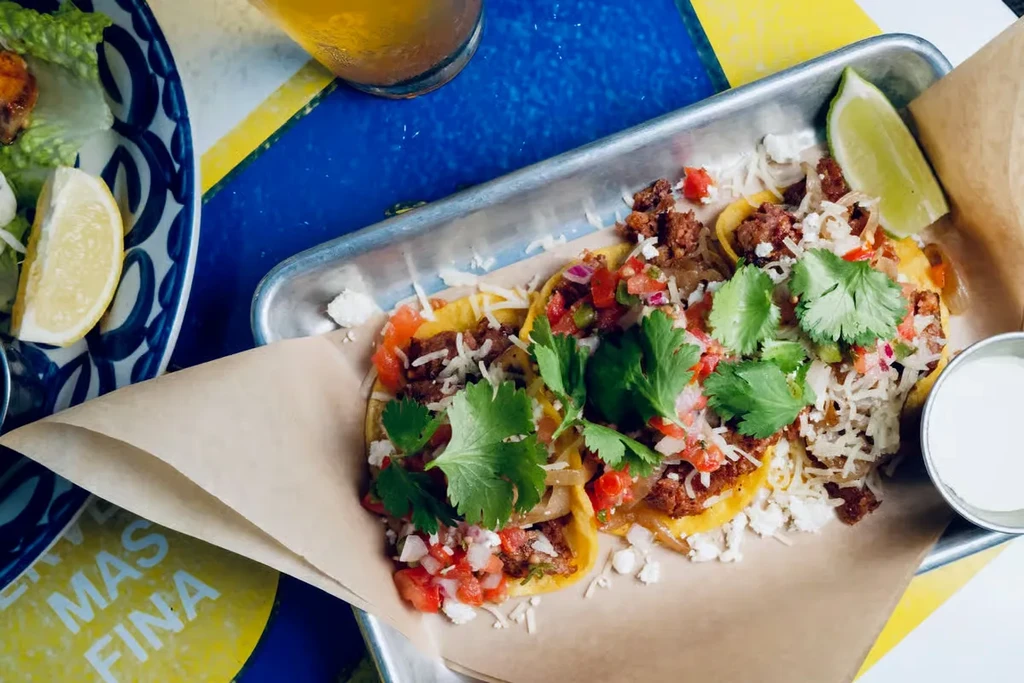 Three street tacos with toppings, sauce, and lime on a colorful table.