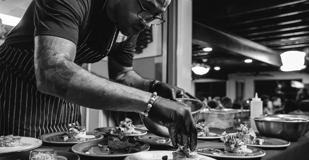 Chef wearing glasses and gloves meticulously plating dishes in a kitchen.