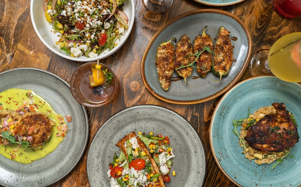 Overhead view of a wooden table with five plates of food and two cocktails.