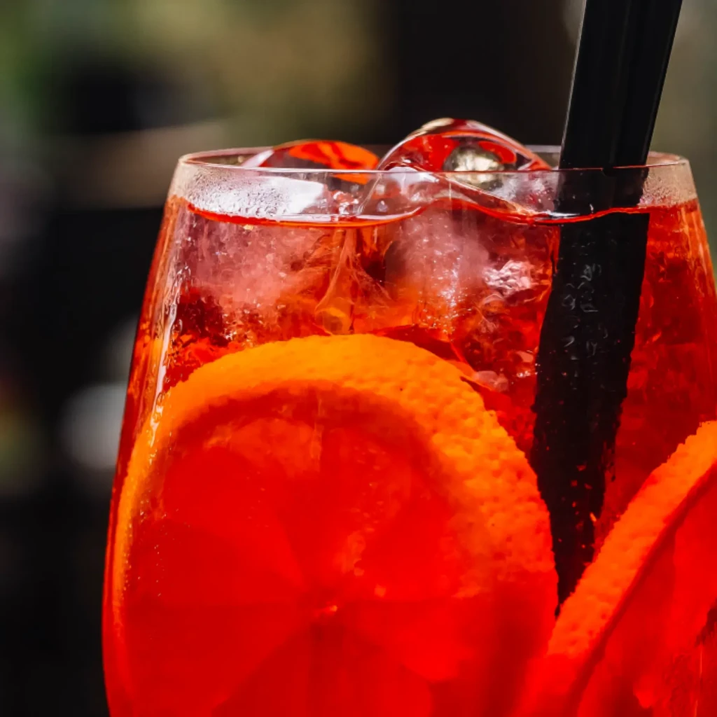 Vibrant red-orange cocktail with ice, orange slices, and a black straw.