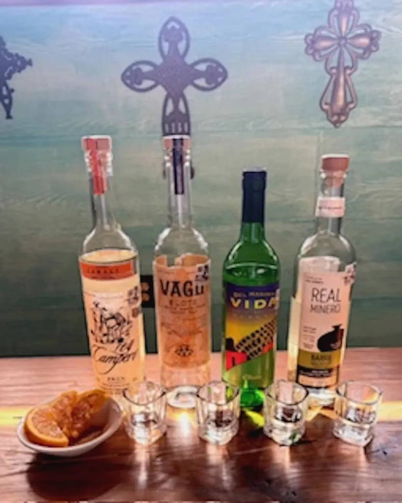 Four bottles of spirits, shot glasses, and orange slices on wood.