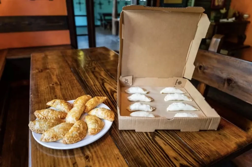 Fried pastries on plate, uncooked pastries in box on wooden table.