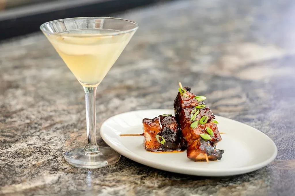 Martini and glazed pork belly skewers on a white plate.