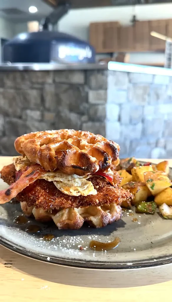 Chicken and waffle dish with a fried egg, bacon, and roasted potatoes.