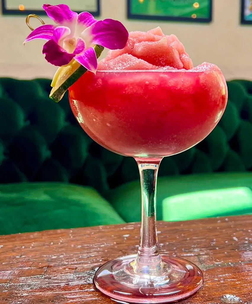 Red frozen drink in a stemmed glass, garnished with orchid and lime.