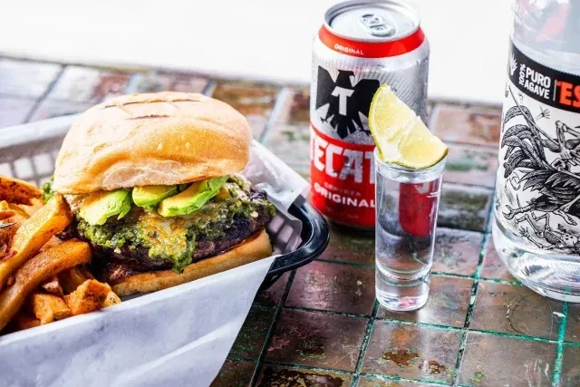 Burger, fries, Tecate beer, shot, and spirit bottle on a table.