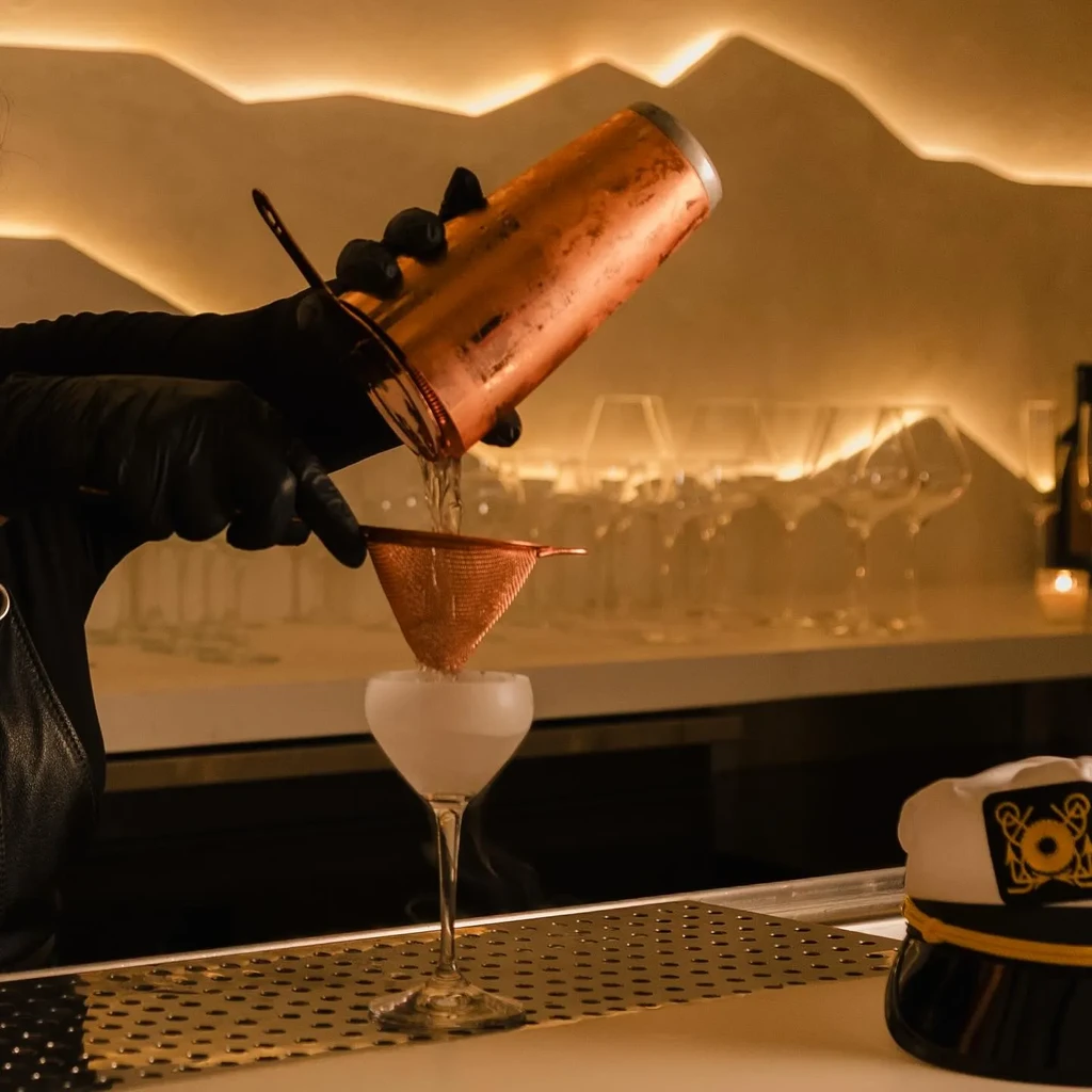 Bartender pouring smoky drink from copper shaker into chilled glass.