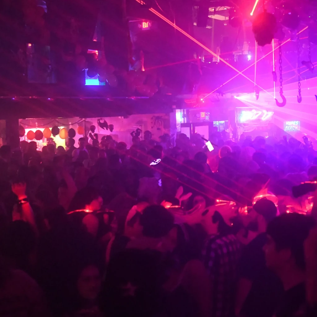 Crowded club interior with purple and pink lights and "HOMIES MERCH" sign.