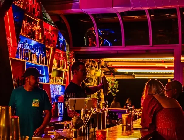 Bartenders work at a neon-lit bar with colorful liquor bottles.