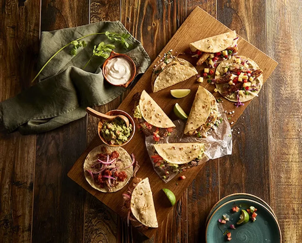 Overhead shot of various tacos, dips, and lime wedges on a wooden table.