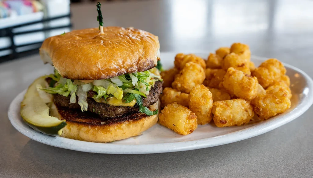 Double cheeseburger with pickle and tater tots on white plate.