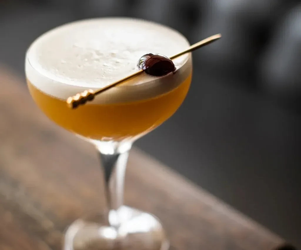 Cocktail in a coupe glass with foam, cherry, and gold pick.