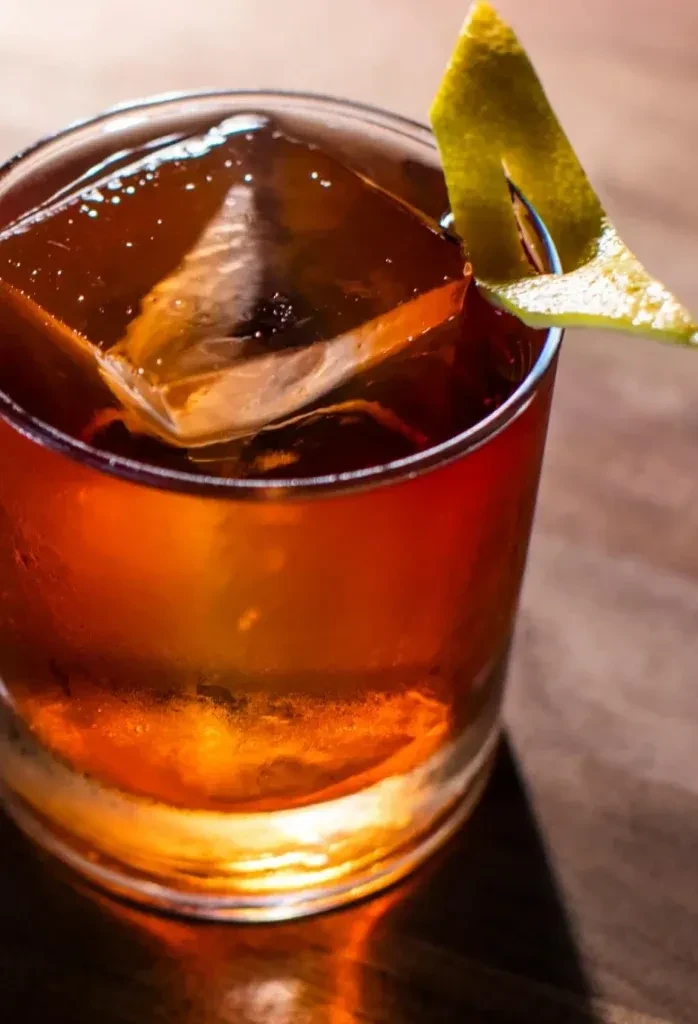 Amber cocktail in a glass with a large ice cube and citrus peel.