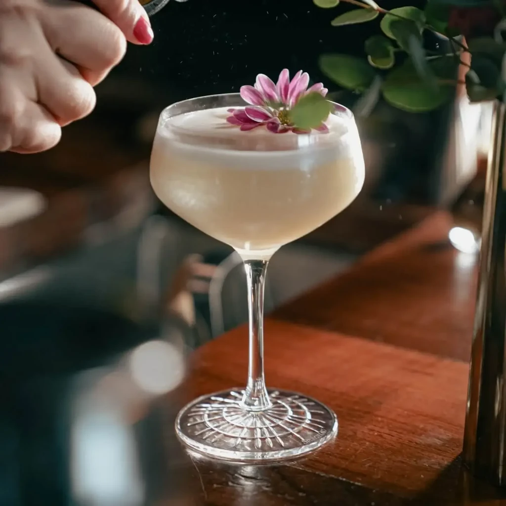 Cocktail in stemmed glass, garnished with flower, hand sprinkles zest.
