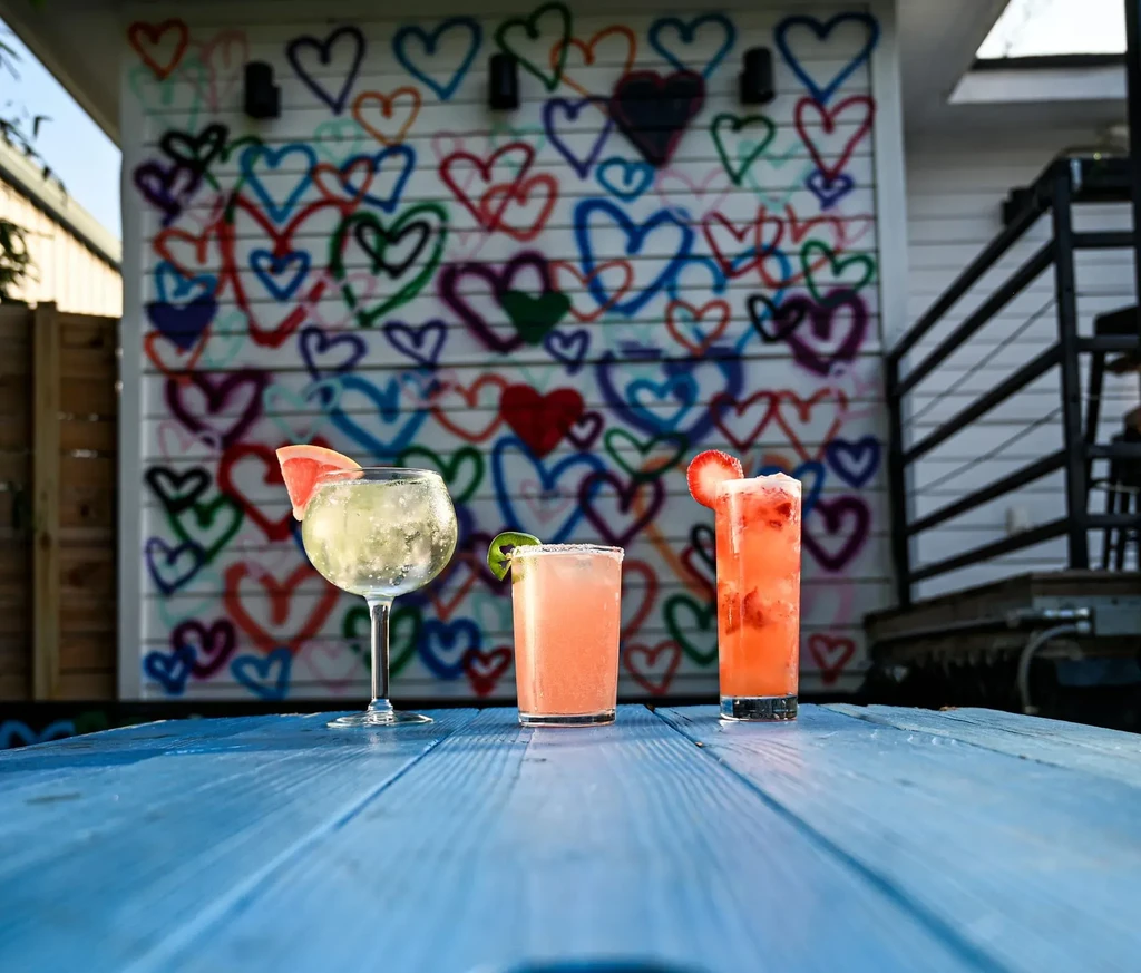 Three colorful cocktails on a blue table with a heart-adorned wall.