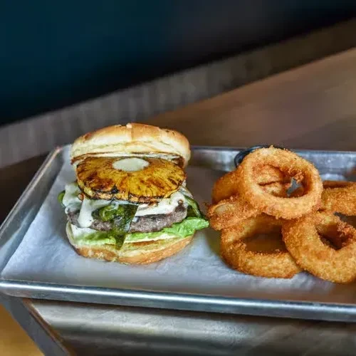 Gourmet burger with pineapple and onion rings on a metal tray.