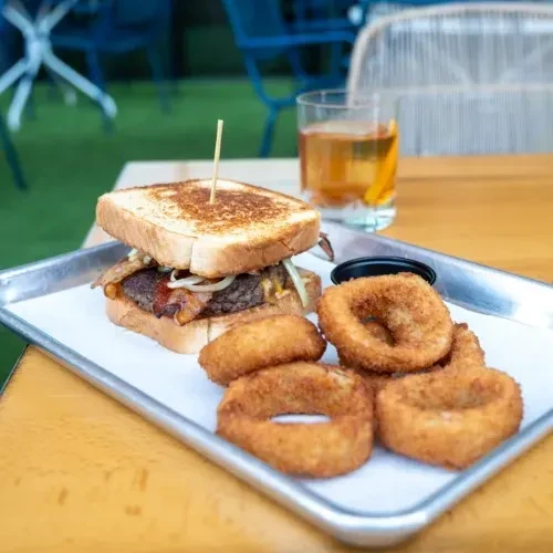Burger or sandwich with onion rings and dipping sauce on a tray.