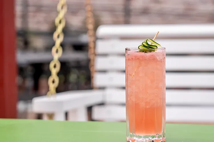 Pink frothy drink with a cucumber garnish on a green table.