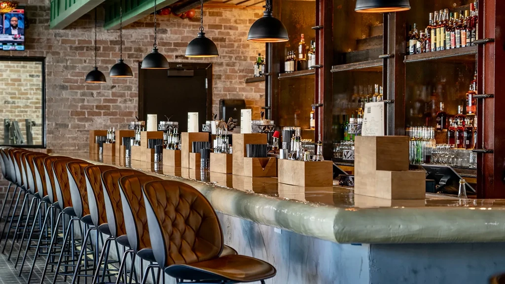 Empty bar counter with stools, liquor bottles, and brick wall.