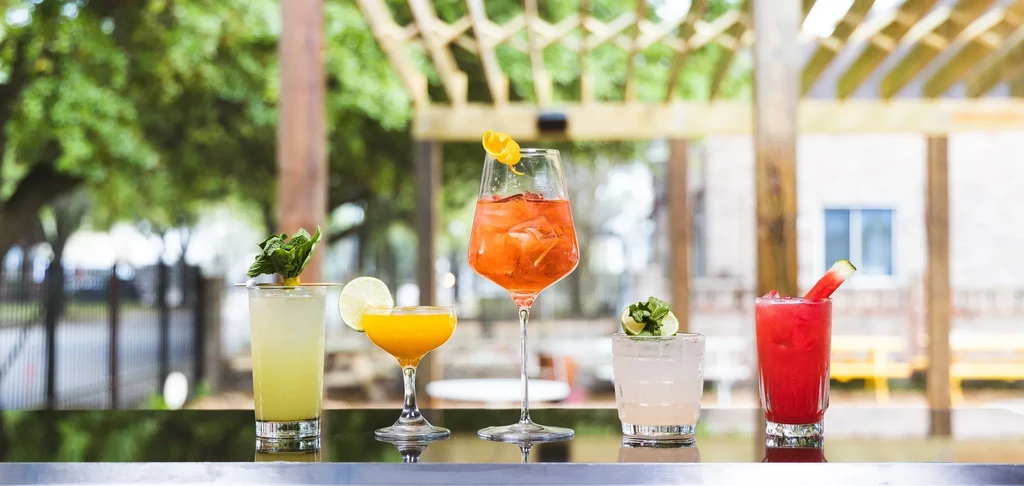 Five colorful cocktails lined up on a reflective bar counter outdoors.