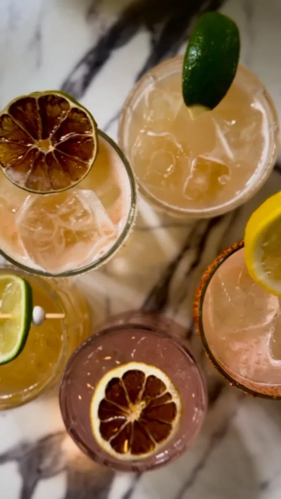 Overhead shot of five cocktails with citrus garnishes on a marbled surface.