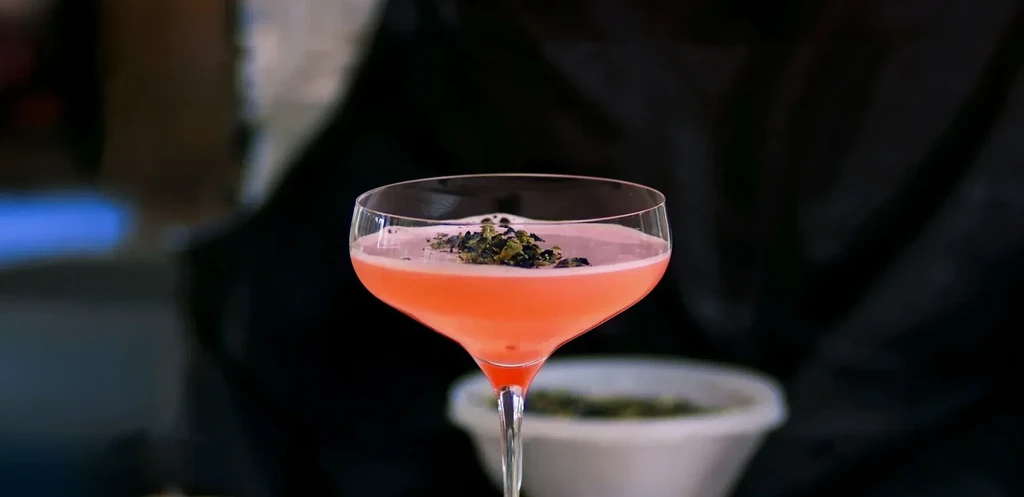 Pink cocktail in a coupe glass with dried herb garnish.