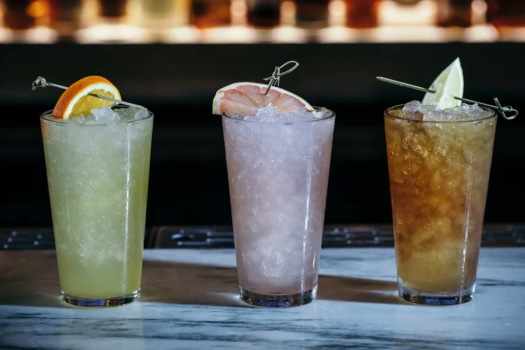 Three iced cocktails garnished with citrus slices on a bar.