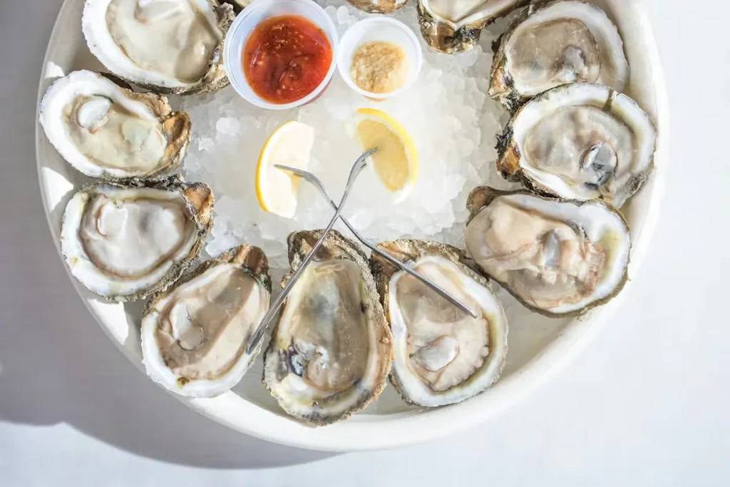 A dozen shucked oysters on ice with sauces, lemon, and forks.