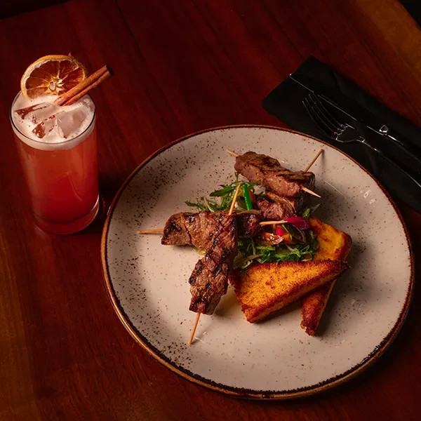 Grilled meat skewers, salad, and cocktail on a dark wooden table.