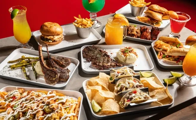 Table with varied food: lamb chops, steaks, burgers, sandwiches, tacos, drinks.