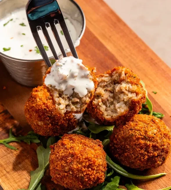 Fried arancini on a wooden board with sauce and greens.
