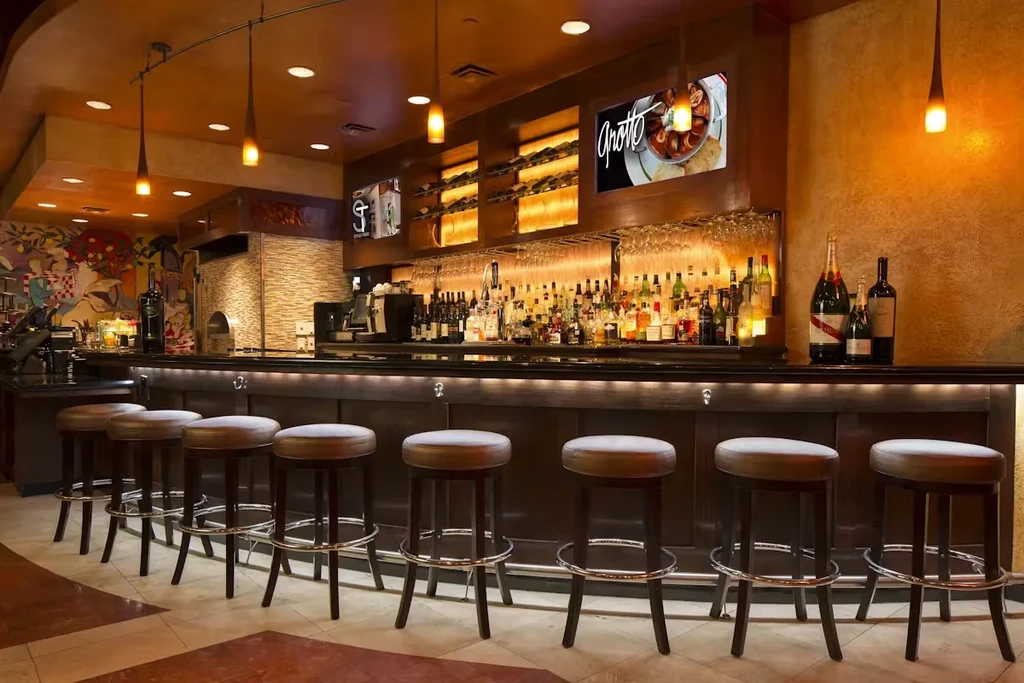 Empty restaurant bar with stools, liquor bottles, and two illuminated screens.