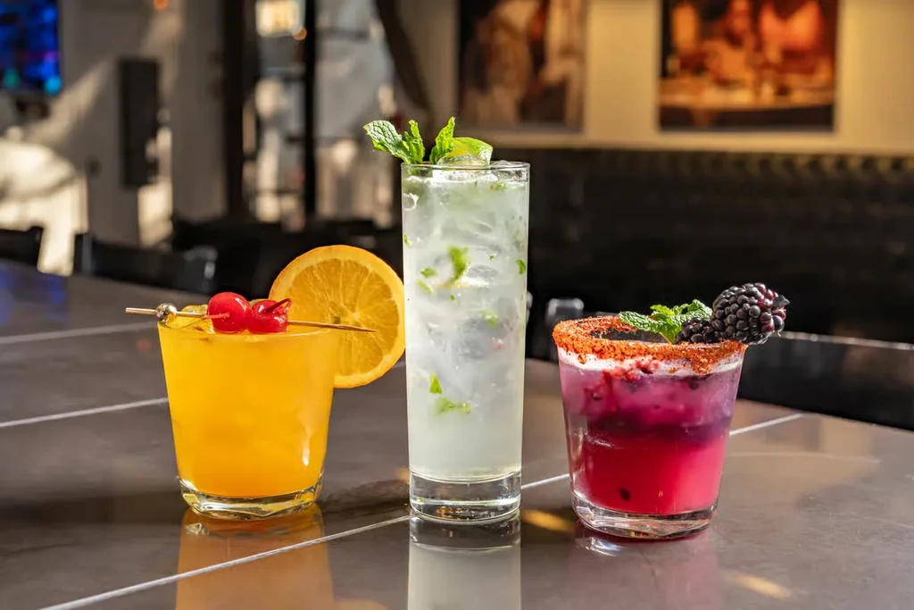 Three cocktails on a dark bar: orange, mojito, and purple with blackberry.