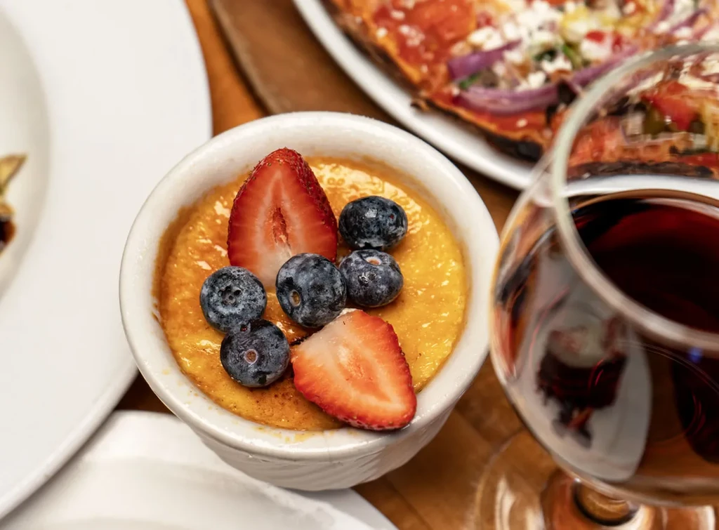Dessert ramekin with fruit, wine glass, and pizza slice on a table.