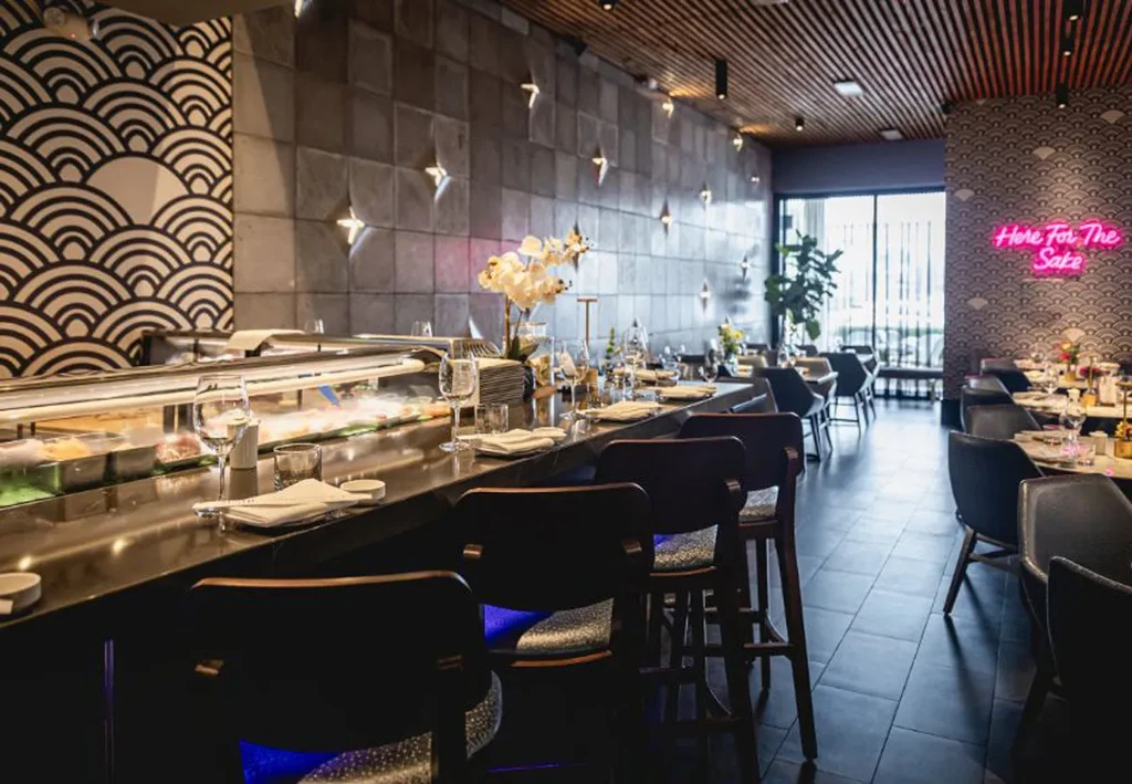 Modern restaurant interior with a bar and "Here For The Sake" sign.