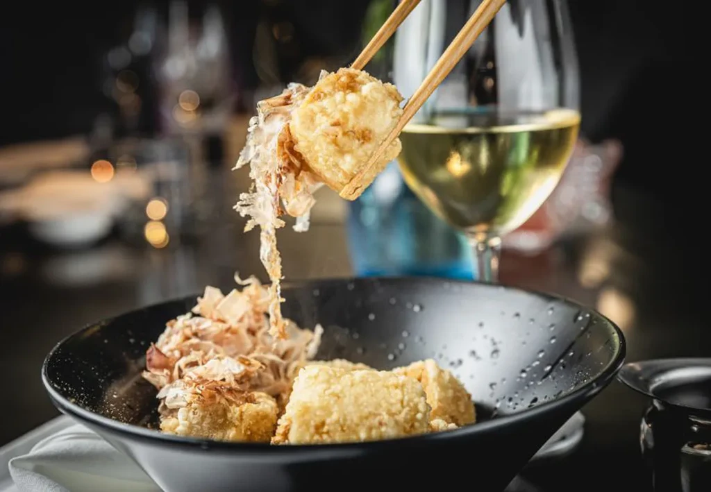 Fried food with flaky toppings, held by chopsticks over a black bowl.