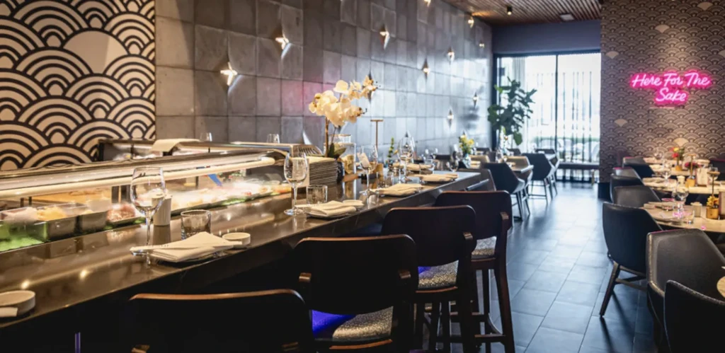 Modern sushi bar with geometric patterns and "Here For The Sake" sign.