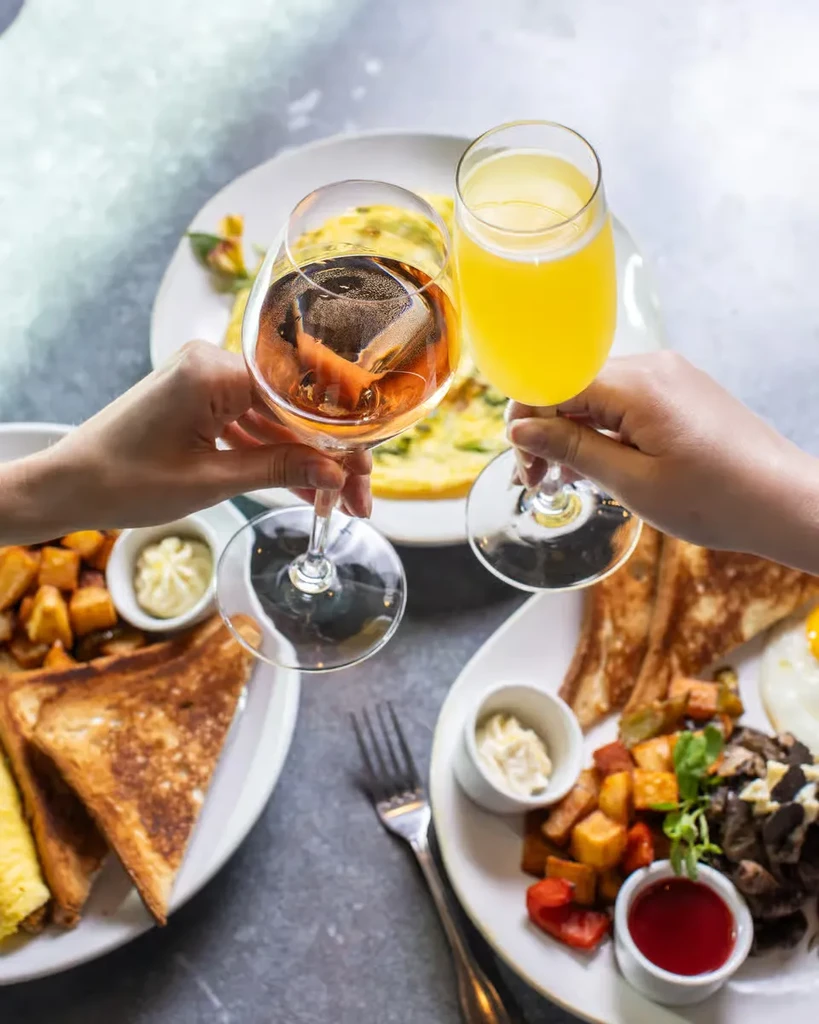 Two people toasting drinks over a table of breakfast food.