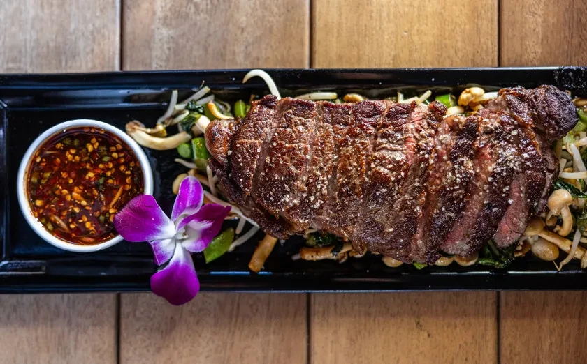 Well-plated sliced steak with vegetables, dipping sauce, and orchid bloom.