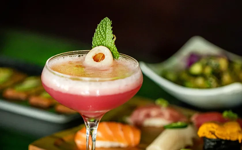 Pinkish-red cocktail with mint and lychee, blurred sushi background.