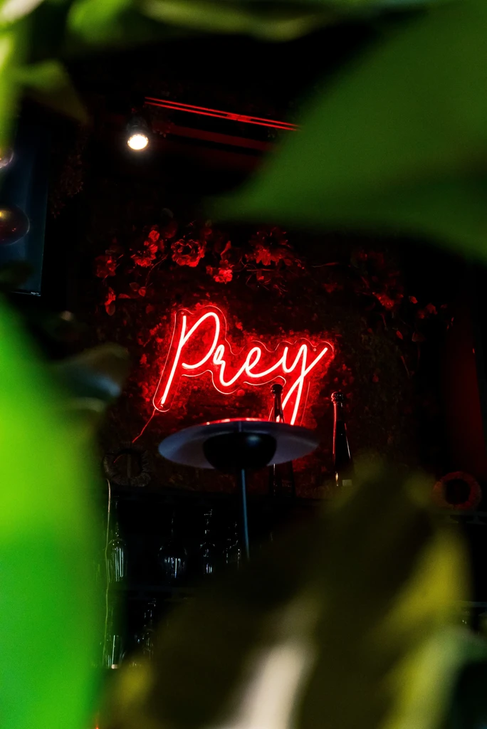 Red neon "Prey" sign behind out-of-focus green leaves.