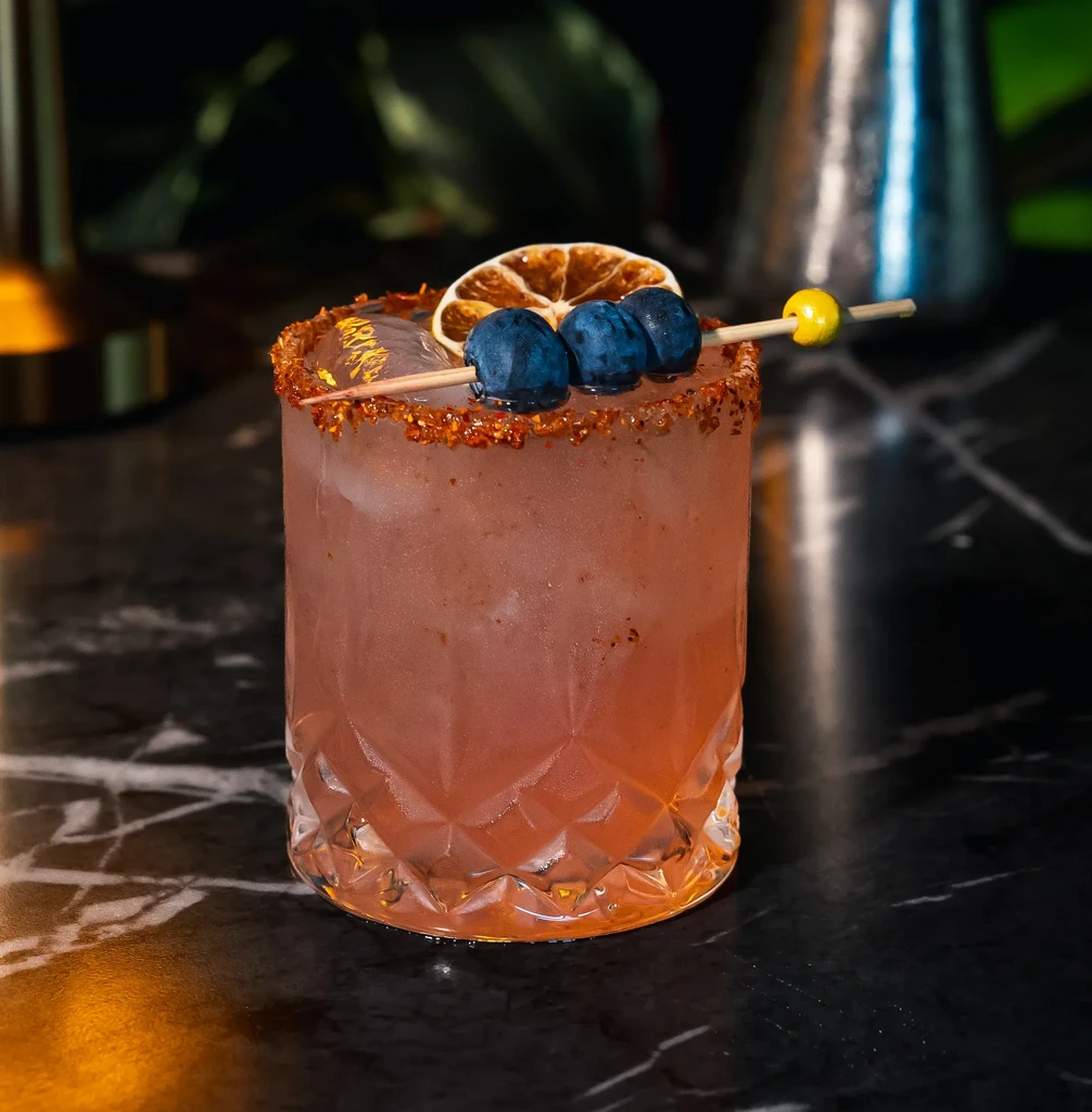 Cocktail in faceted glass with berries, citrus, and spiced rim.