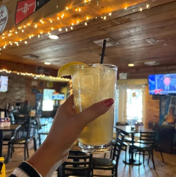 Hand holding a lemon-garnished drink in a dimly lit rustic bar.