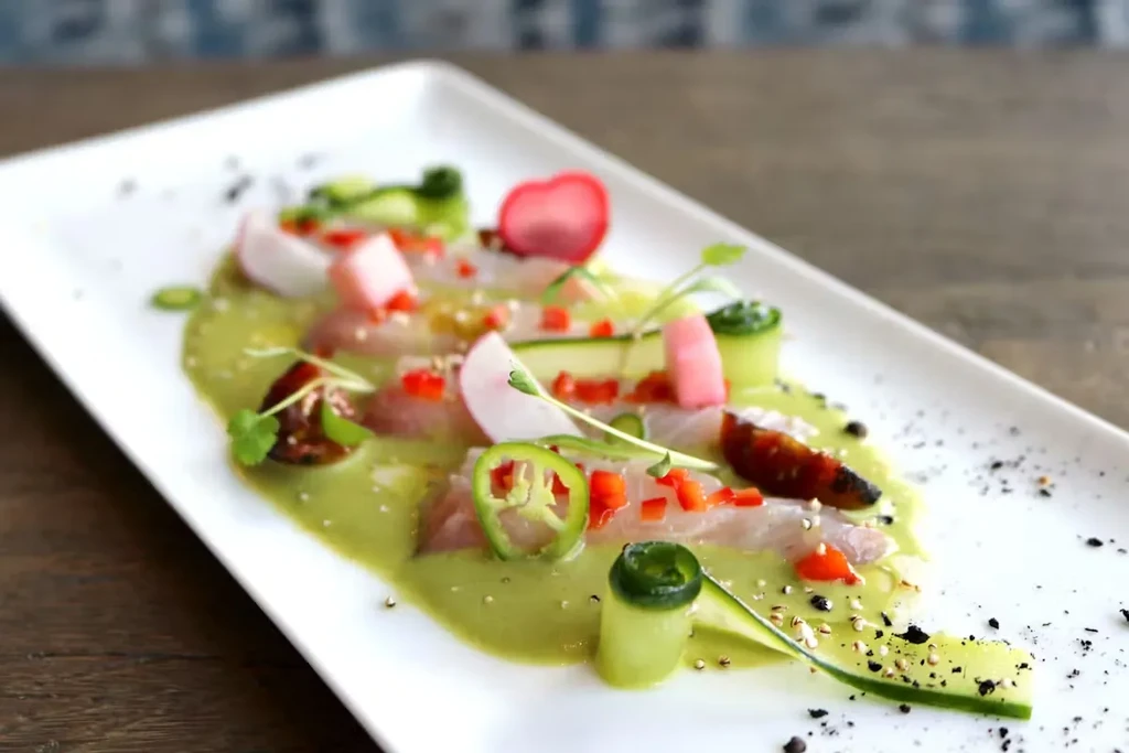 Plated fish dish with various colorful garnishes and green sauce.