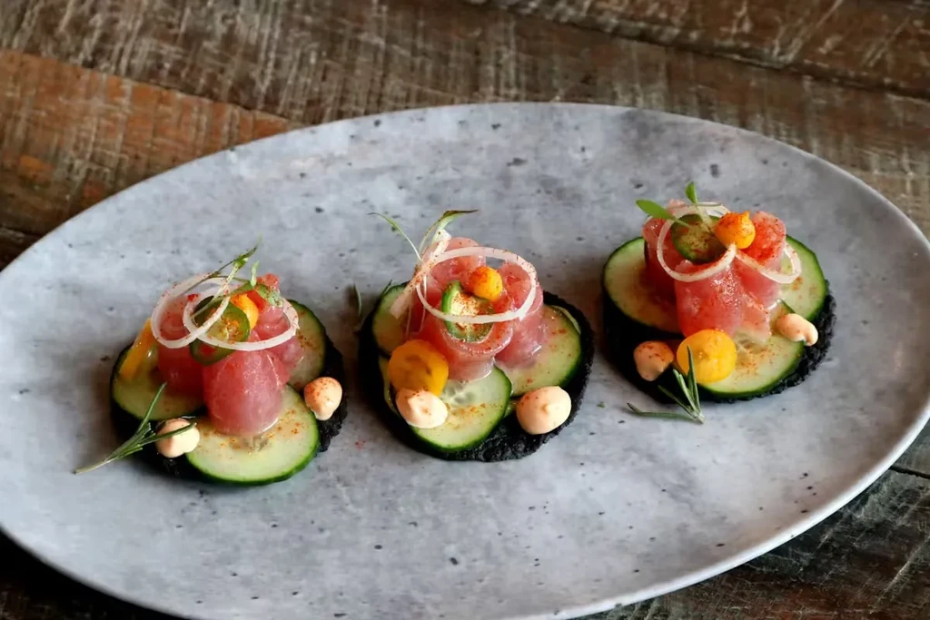 Three appetizers with fish, cucumber, and various garnishes on a plate.