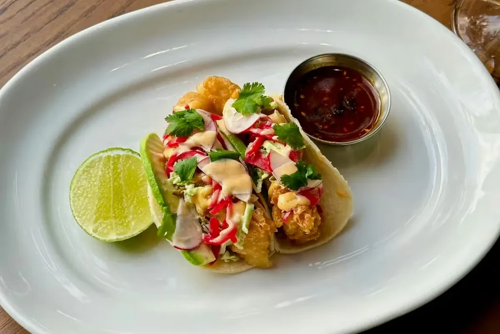 Two fish tacos with toppings, lime, and sauce on a white plate.