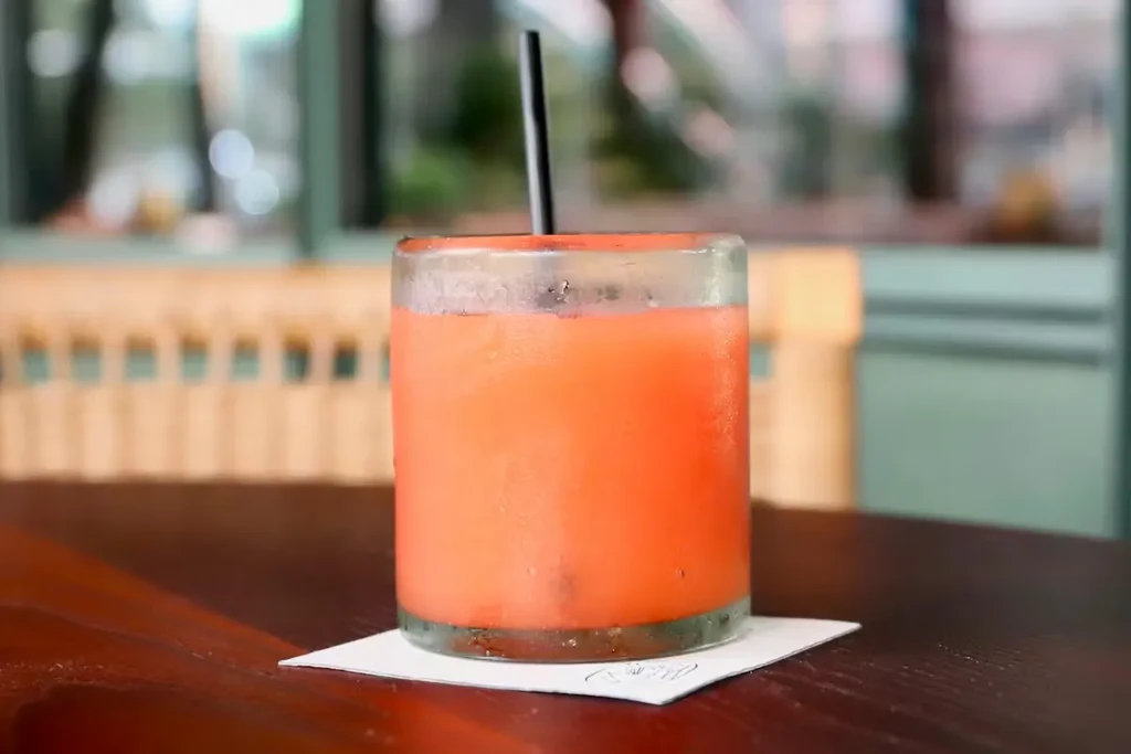 Frosted glass with reddish-orange drink, black straw, on wooden table.